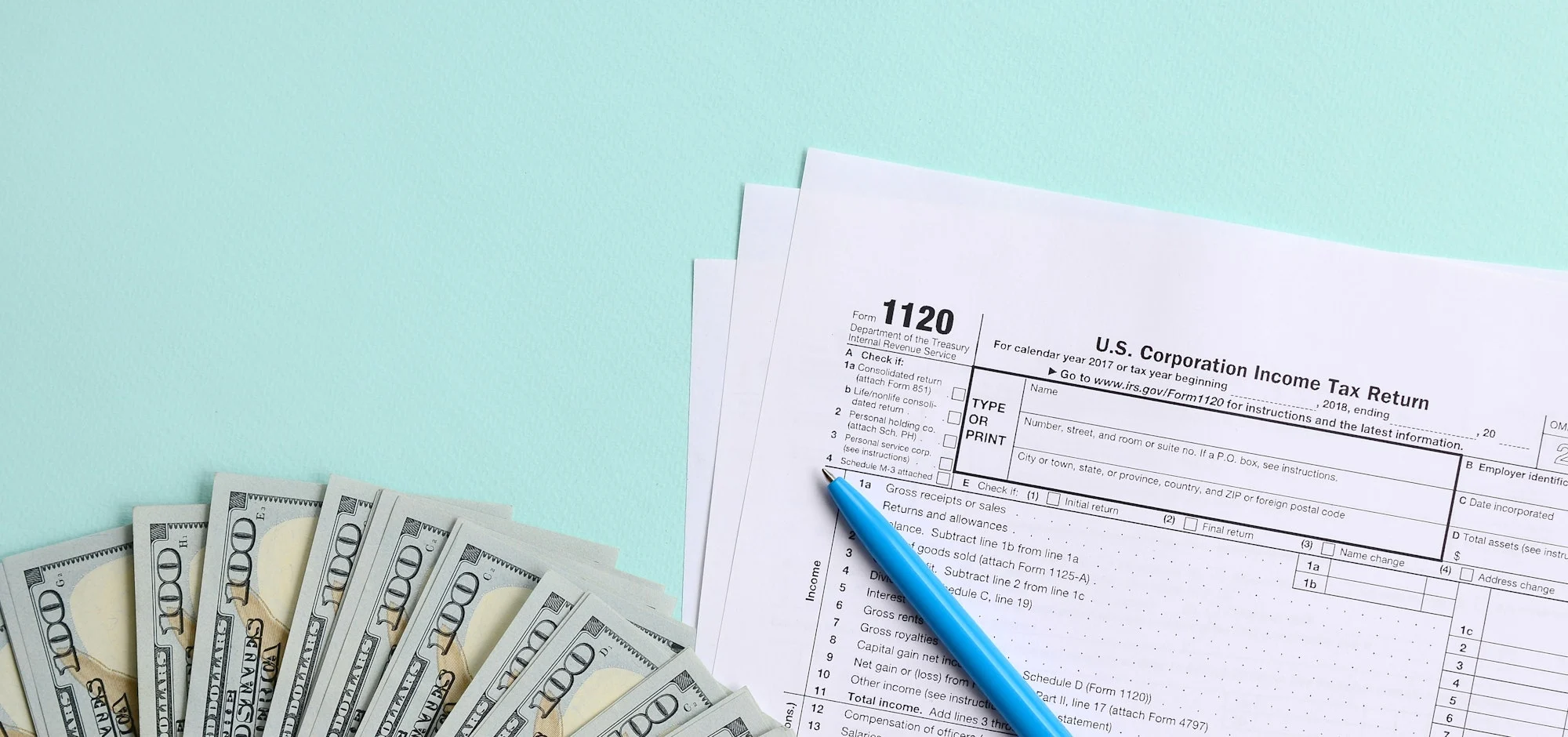 1120 tax form lies near hundred dollar bills and blue pen on a light blue background